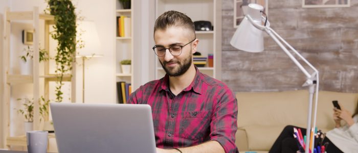 handsome-man-laughing-while-working-laptop-living-room-girlfriend-background-talks-phone
